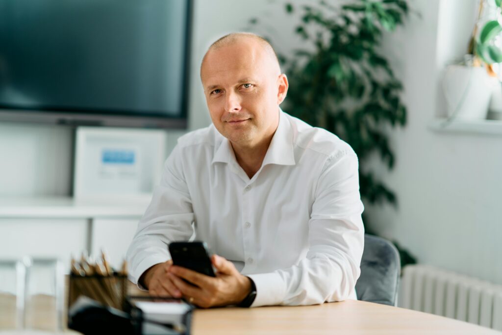 Businessman in a white shirt sitting in a modern office, holding a smartphone, and looking confident.